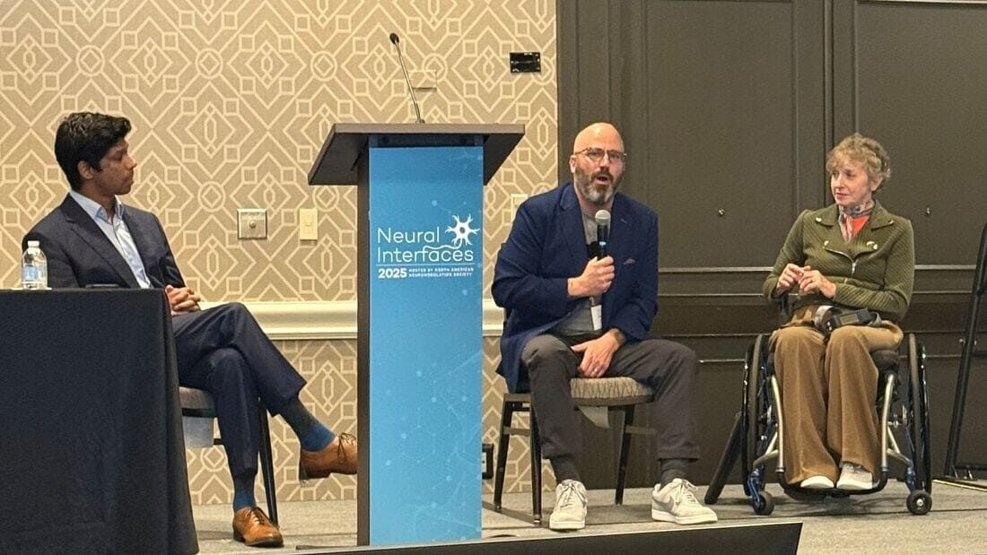 Three people participate in a panel discussion; one speaks into a microphone, another sits in a wheelchair, and the third listens. A podium with a Neural Interfaces 2023 sign is placed at the center.