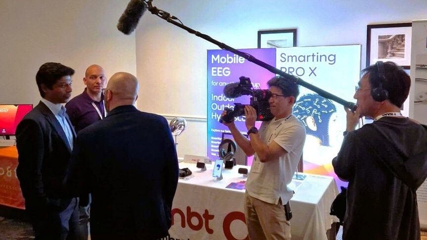 A camera crew films two men talking at a tech expo booth displaying Smarting PRO X and Mobile EEG signs. A boom microphone is held overhead, and various electronic devices are on the table.