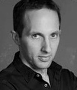 A man with short dark hair wearing a dark shirt looks seriously at the camera. The image is black and white with a plain background.