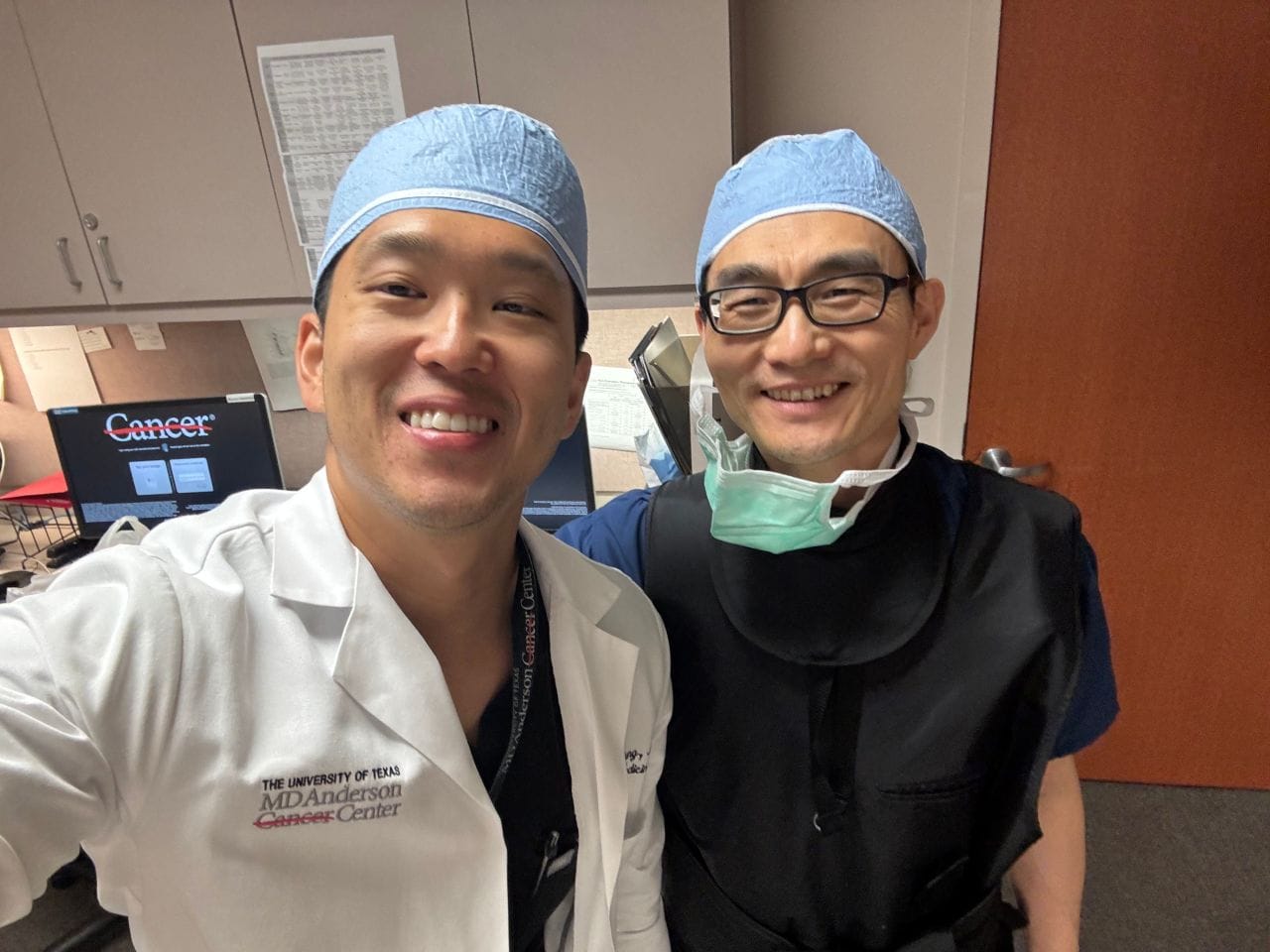 Two medical professionals in scrubs and surgical caps smile for a selfie in a hospital or clinic setting. One wears a white coat with MD Anderson Cancer Center and the other wears a protective vest and mask around his neck.