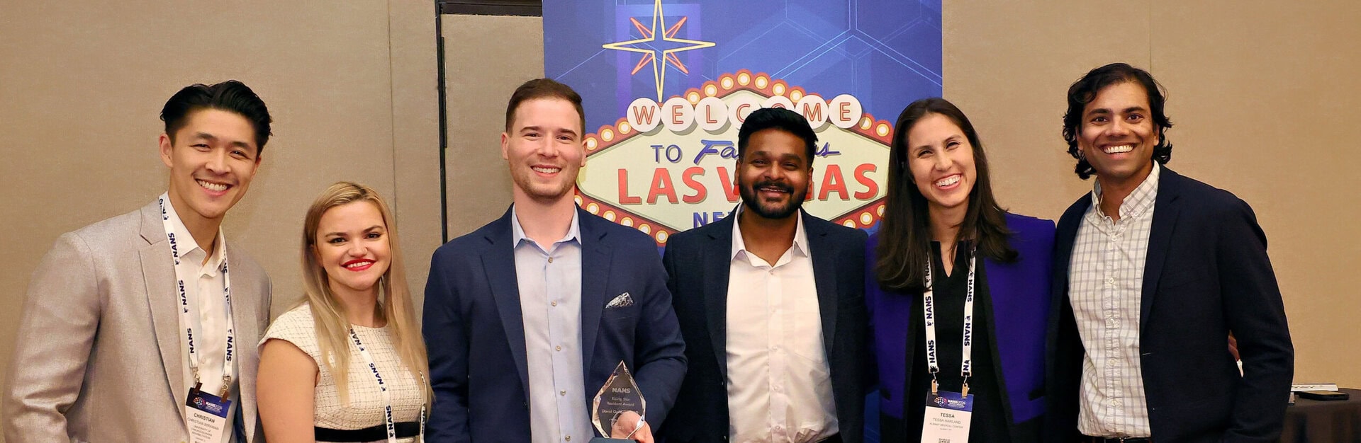Six people in business attire smile and pose together in front of a Welcome to Fabulous Las Vegas sign at an indoor event. One person holds a glass award.