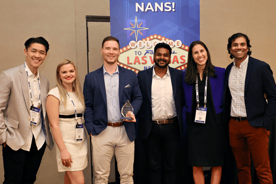 Six people stand smiling in front of a Las Vegas-themed sign at what appears to be a professional event. One person holds a small trophy. Most wear business or business-casual attire and event badges.