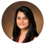 A woman with long black hair and a maroon sleeveless top smiles at the camera against a brown studio background.