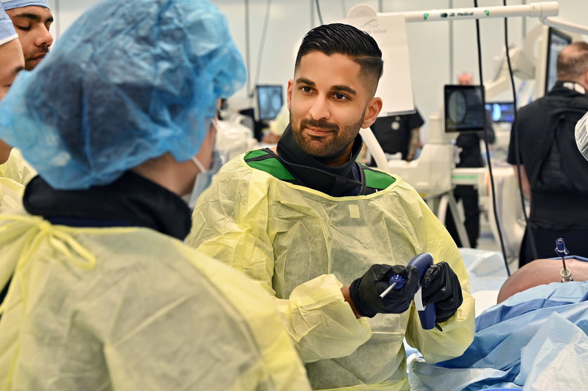 A medical professional in yellow gowns and gloves holds an instrument, engaging with a colleague in blue scrubs in a clinical setting with monitors in the background.