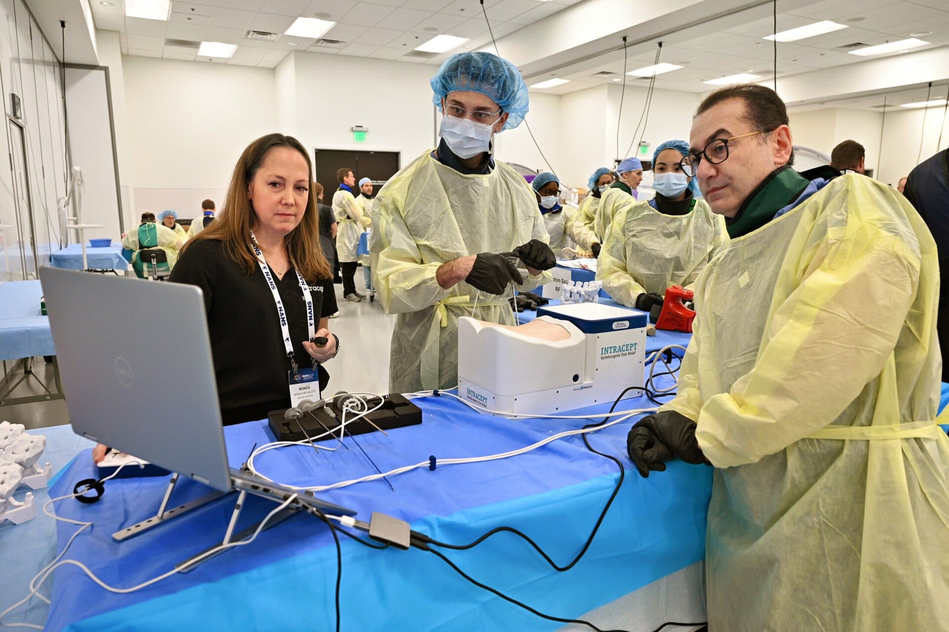 Four people in medical gowns and masks stand around a table with a laptop and equipment in a bright lab, appearing to be in discussion or at a workshop.