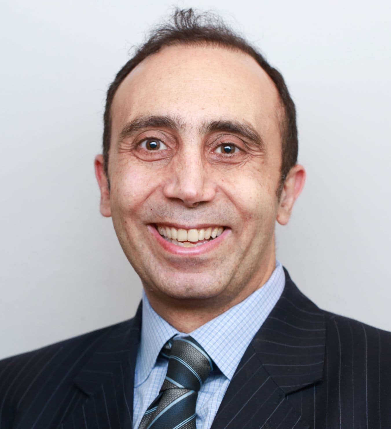 Smiling man with short dark hair wearing a pinstripe suit, blue checked shirt, and striped tie, posing in front of a plain light background.