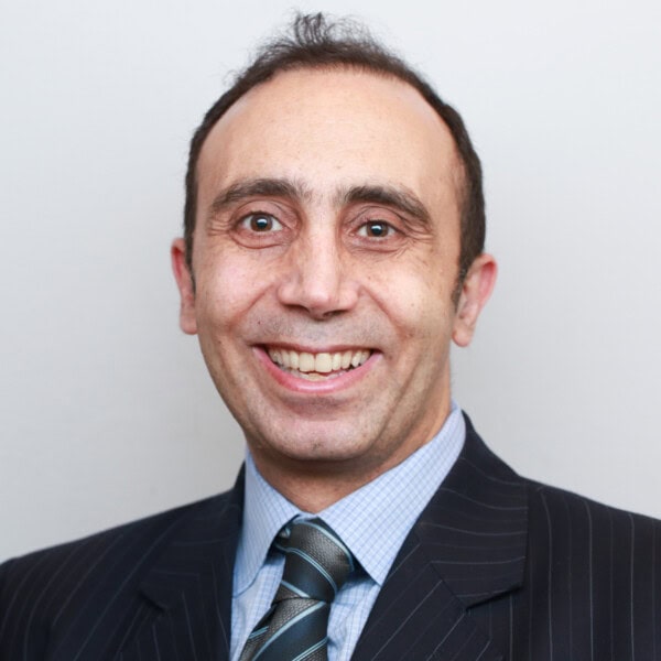 A man in a dark pinstripe suit, blue checkered shirt, and striped tie smiles at the camera against a plain light background.