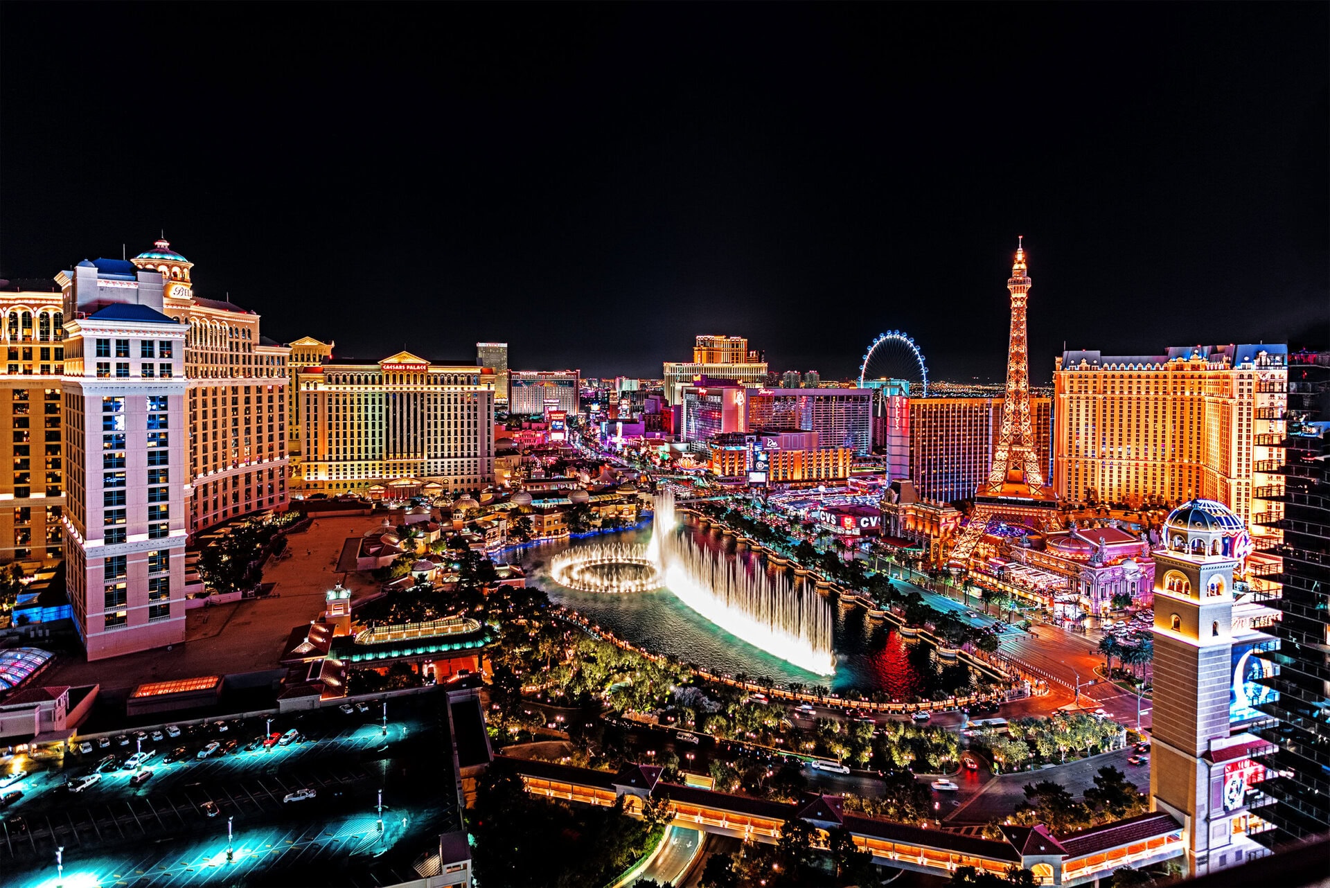 Las Vegas Strip at night, featuring brightly lit hotels, casinos, the Bellagio fountains in mid-display, and a large Ferris wheel in the background.