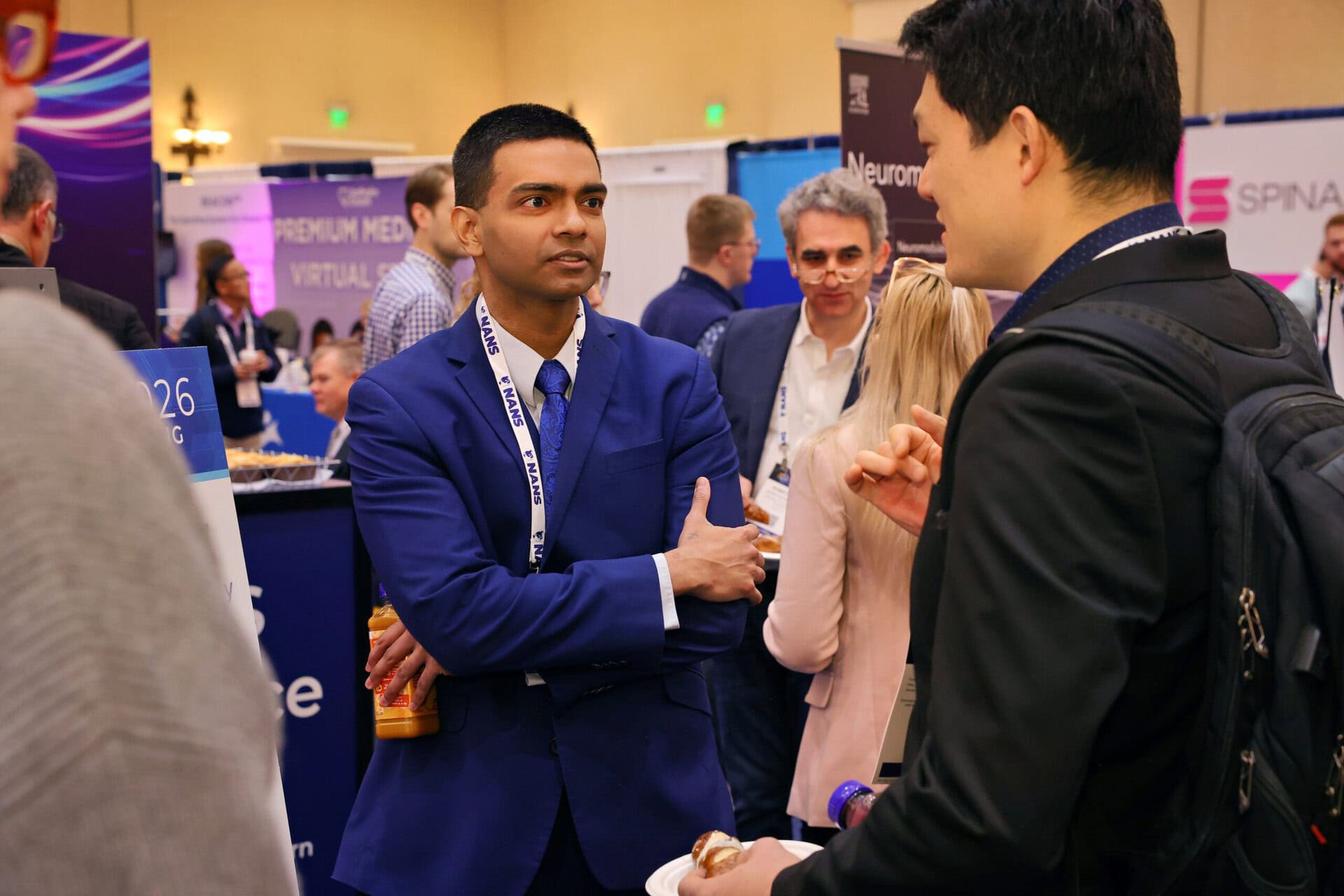 A man in a blue suit holding a drink listens to another man at a busy conference or expo, with booths and attendees visible in the background.