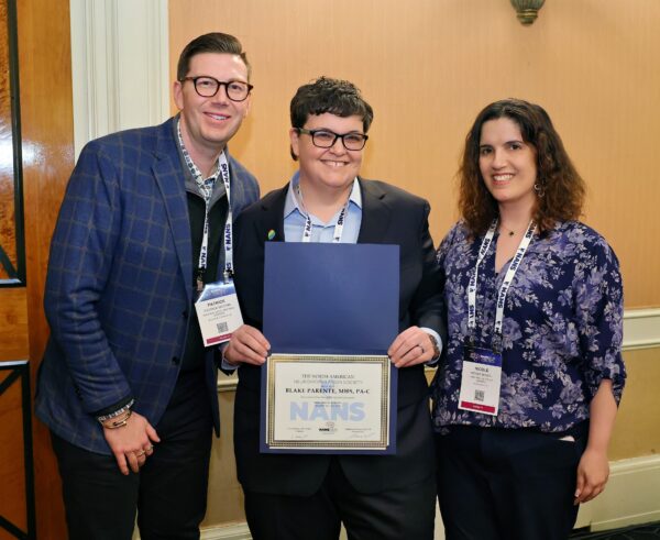 Three people stand together indoors, smiling. The person in the middle holds a certificate with a NANS logo. All are wearing conference badges. Theres a plaque and tan wall in the background.