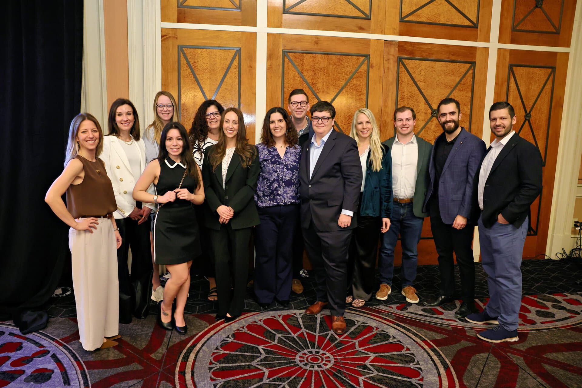 Thirteen people stand together indoors, smiling in business-casual attire, in front of a wood-paneled wall with geometric designs and carpeted flooring.