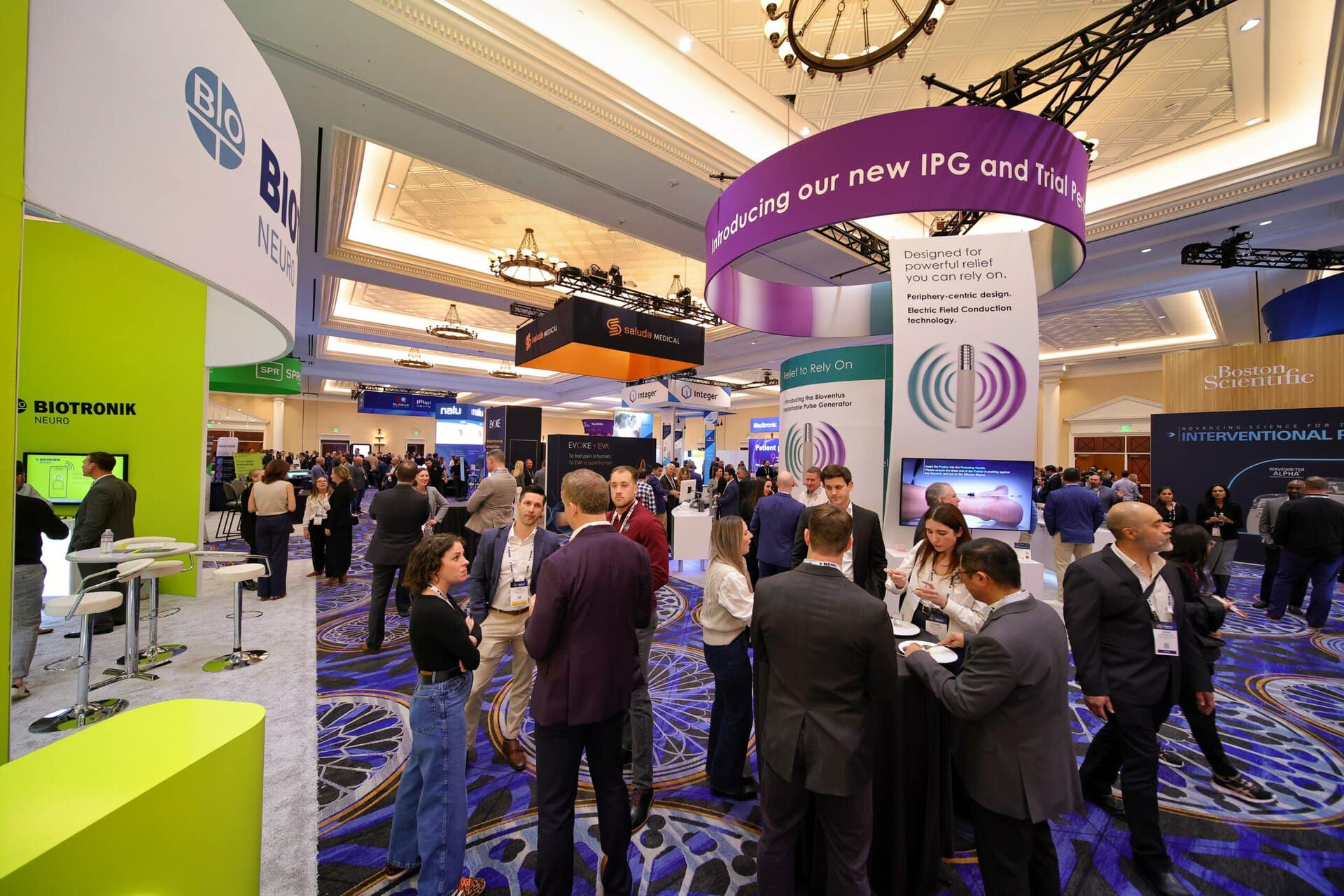 A busy convention hall with people networking among colorful booths and banners. Attendees wear badges and engage in conversations under large hanging signs promoting medical and scientific companies.