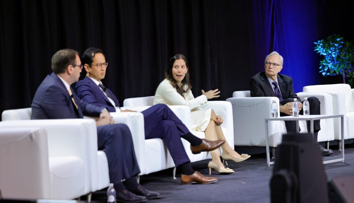 Four people in business attire sit on a stage in white chairs, engaged in a panel discussion. One woman gestures while speaking, and the others listen. A small table with water bottles is in front of them.