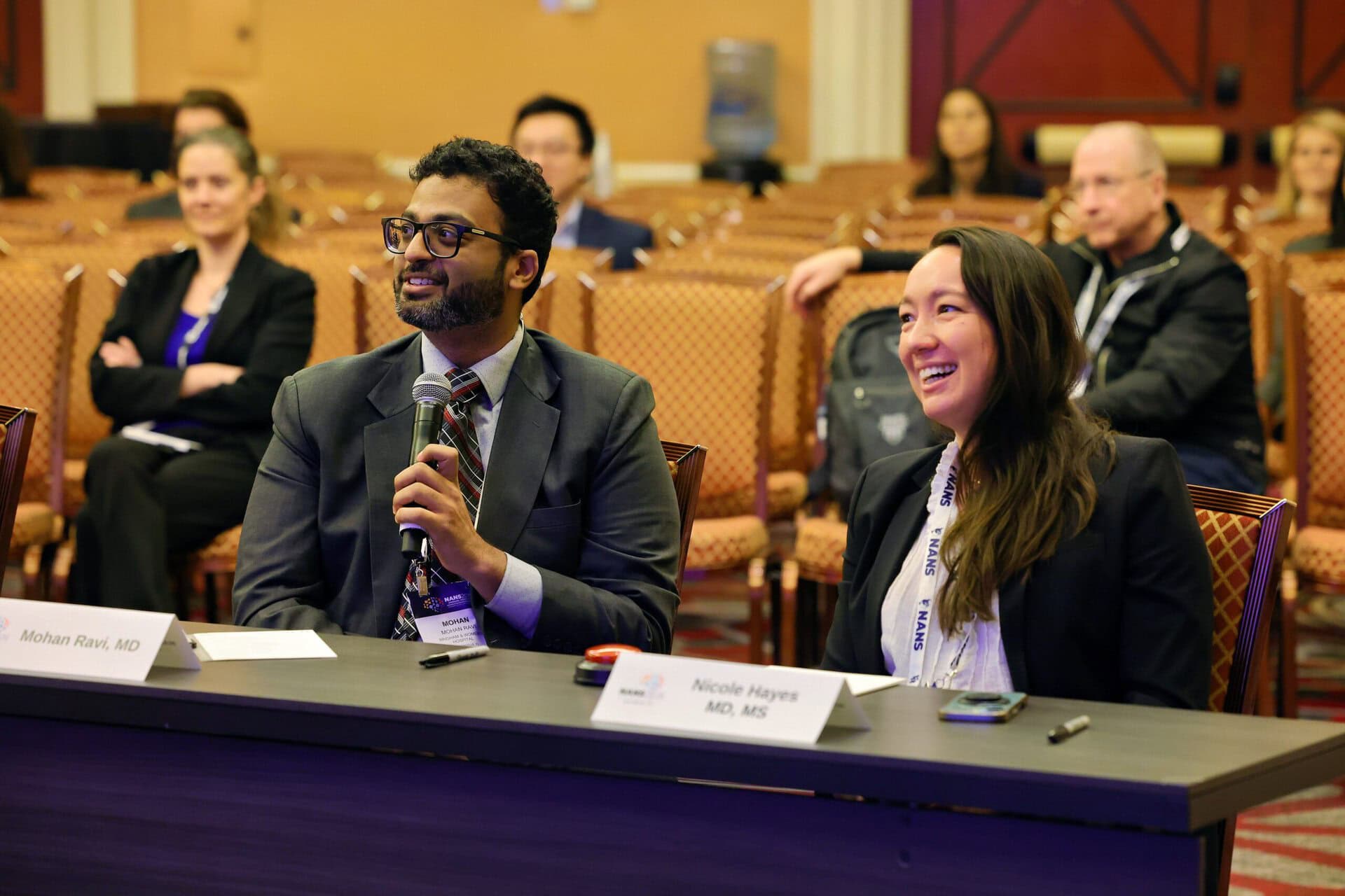 Two people in business attire sit at a panel table, smiling and engaging with the audience. Several attendees are seated in rows behind them, listening attentively.