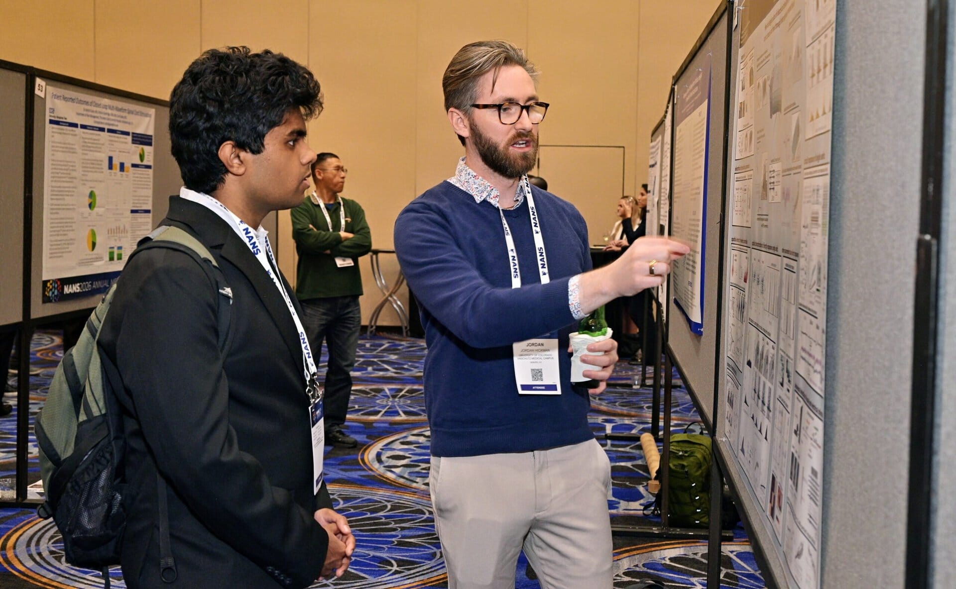 Two men stand in front of a scientific poster at a conference. One points to the poster while explaining, and the other listens attentively. People and other posters are visible in the background.