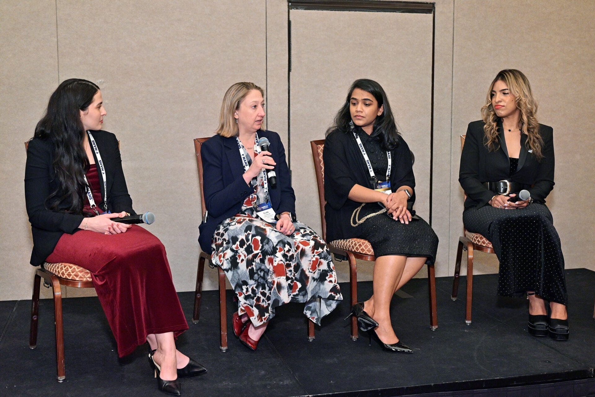 Four women sit on chairs in a panel discussion. One woman speaks into a microphone while the others listen attentively. They are dressed in professional attire and wear conference lanyards.