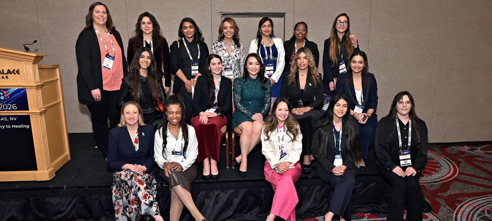 A group of women in professional attire pose together at a conference in a carpeted room, with some seated and others standing. A podium and conference signage are visible on the left side.