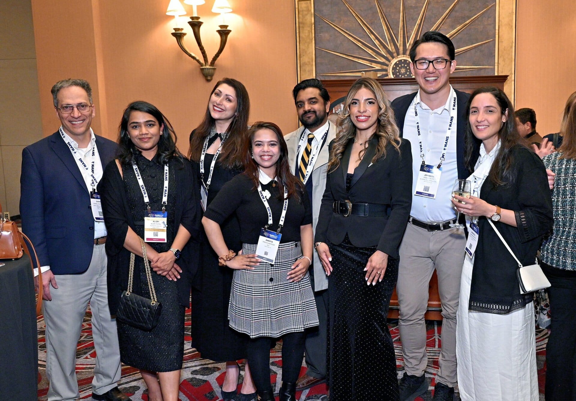 A group of eight people in business attire, smiling and posing together at a conference or networking event. They wear name tags and stand in front of a sunburst wall decoration.