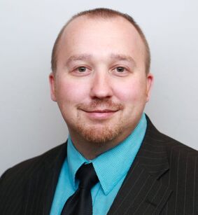 A man with short light brown hair, trimmed beard, and mustache, wearing a black pinstripe suit, teal shirt, and black tie, smiles at the camera against a plain light background.