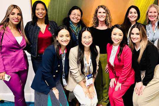 Nine women, dressed in business attire, are smiling and posing together indoors at what appears to be a professional event or conference. Some wear suits and conference badges, standing in front of a colorful backdrop.